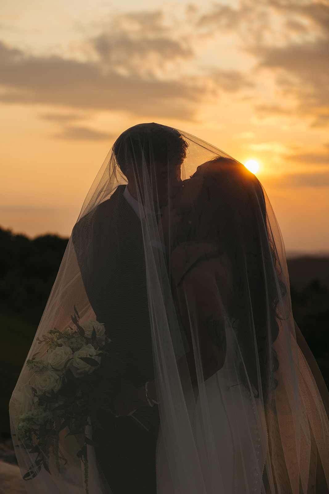Sunset wedding photo of bride and groom under a veil kissing