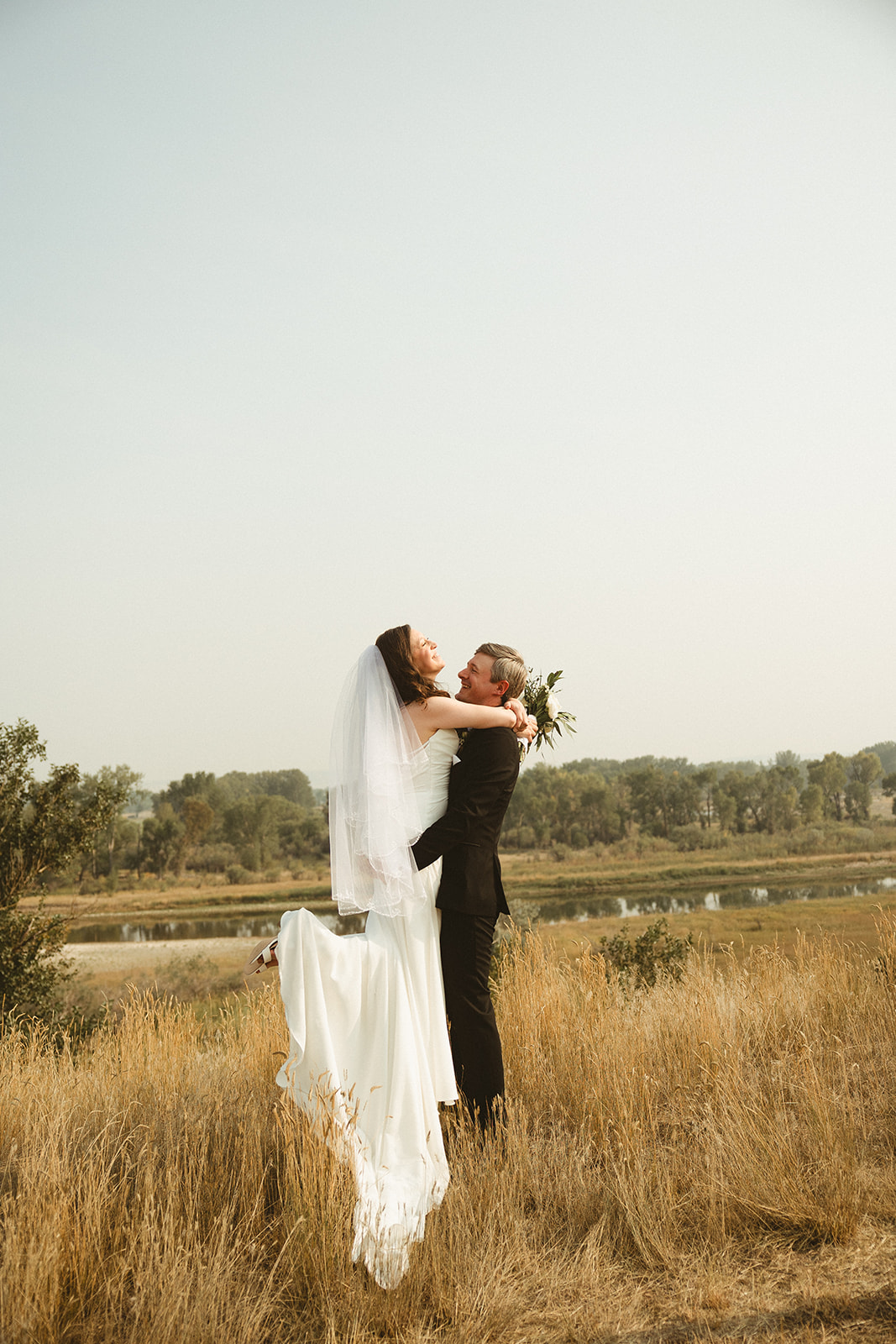 A bride and groom in a field taking wedding photos with the groom lifting her up