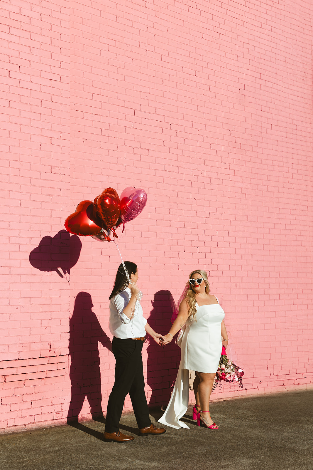 Two brides posing in front of a pink brick wall with heart balloons, perfect for unique wedding ideas
