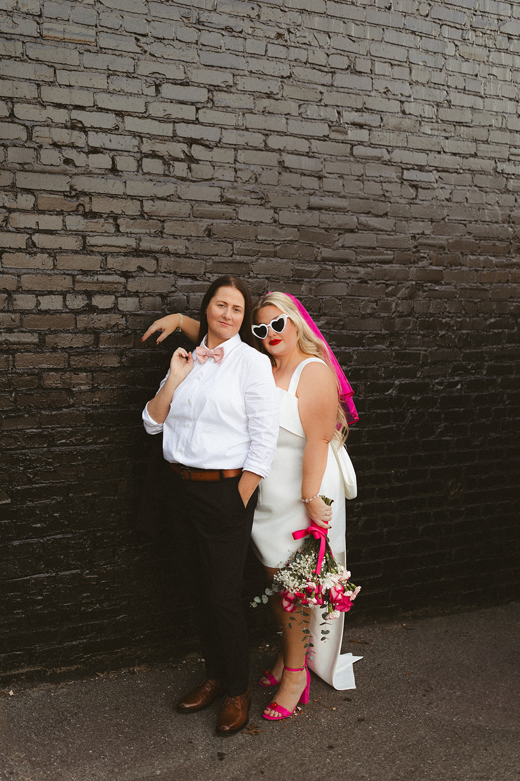 Two brides posing for elopement photos in front of a black brick wall