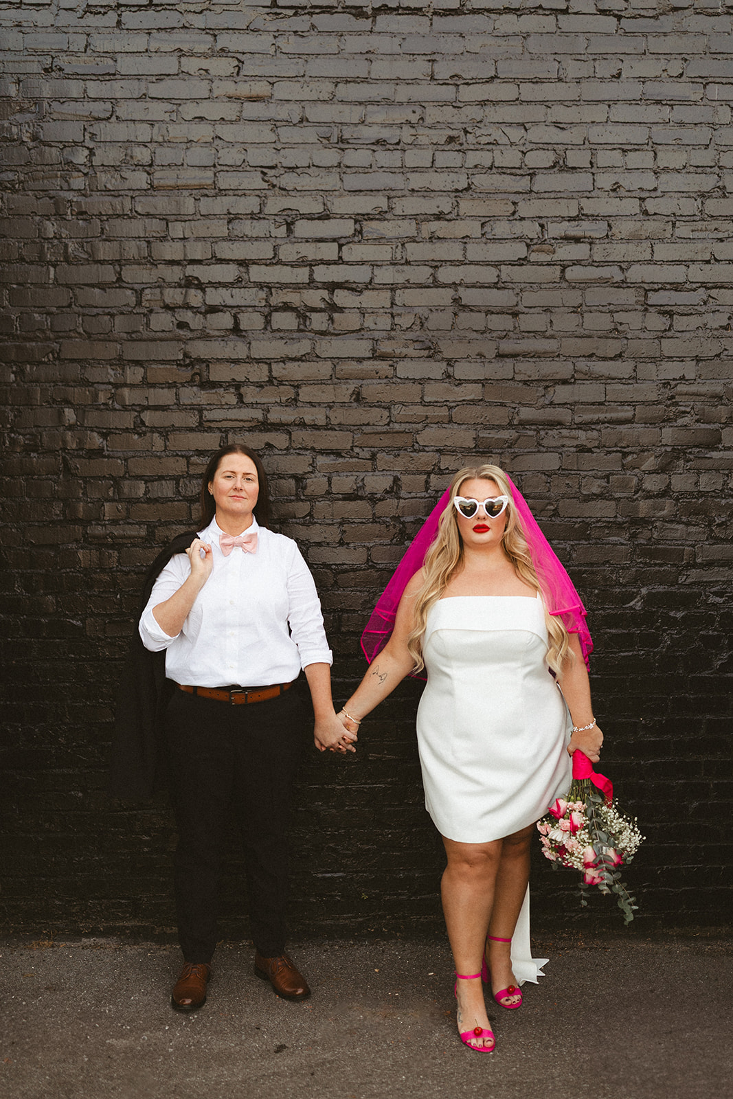 Two brides posing for elopement photos in front of a black brick wall