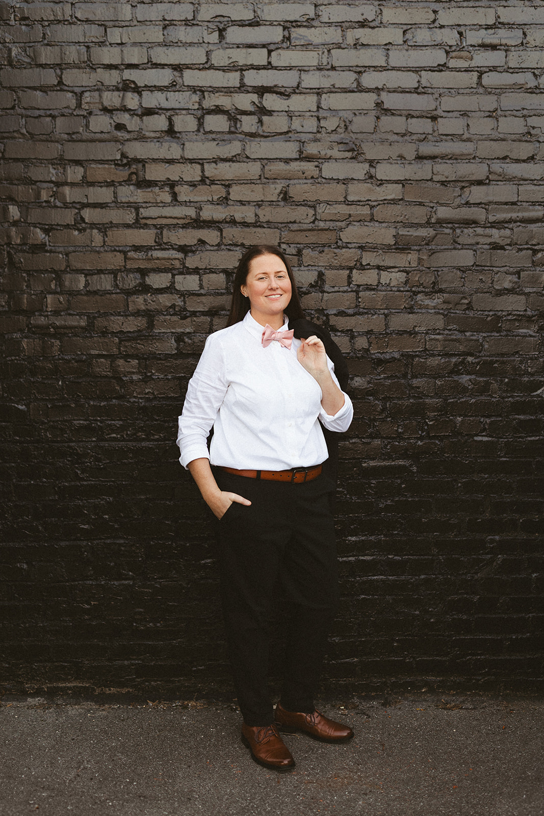 A bride's suit and bowtie for a wedding, while she's posing in front of a black brick wall