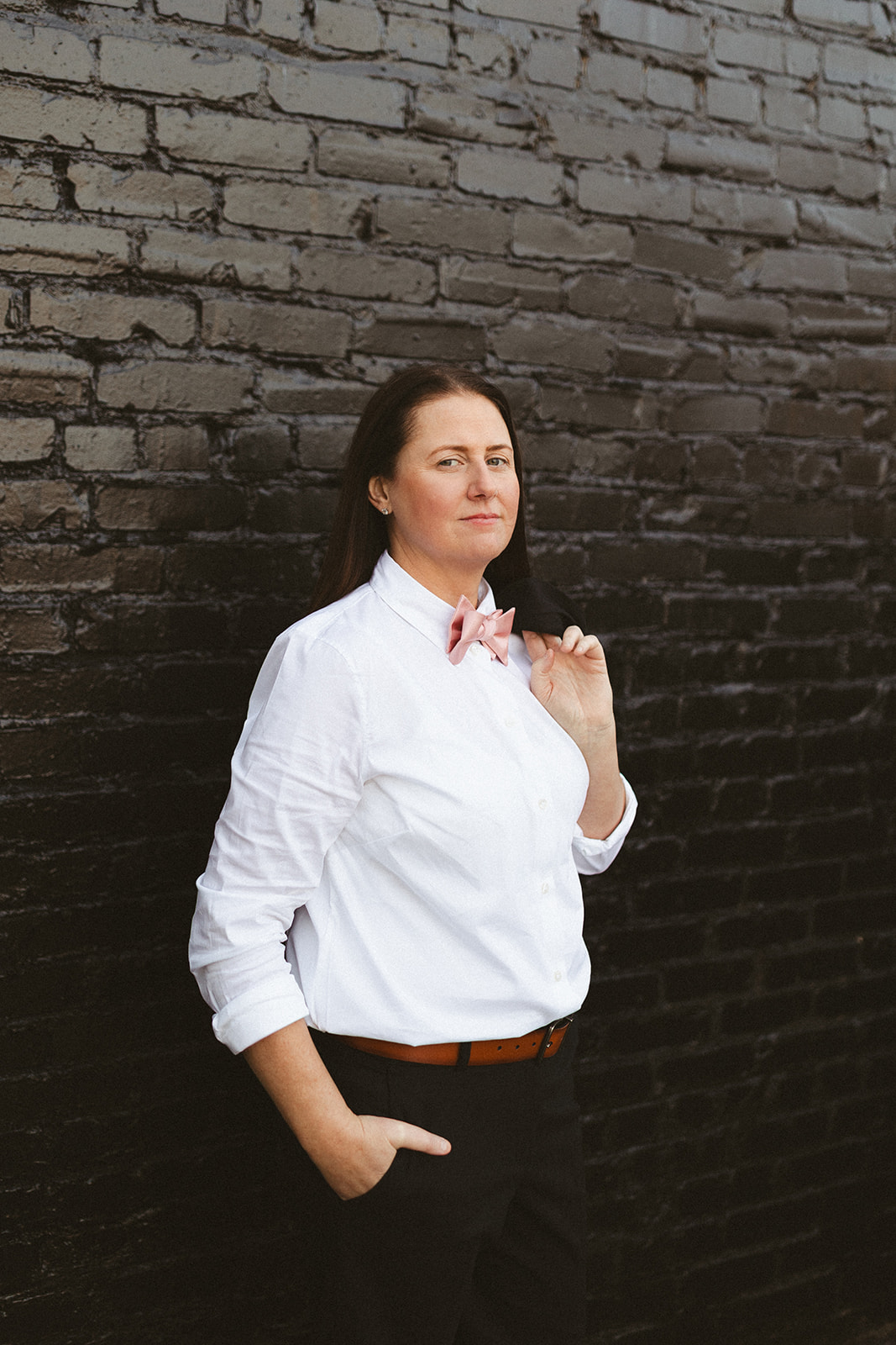 A bride's suit and bowtie for a wedding, while she's posing in front of a black brick wall
