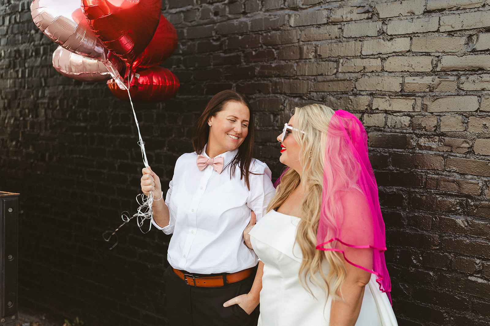 Two brides smiling at each other leaning against a black brick wall