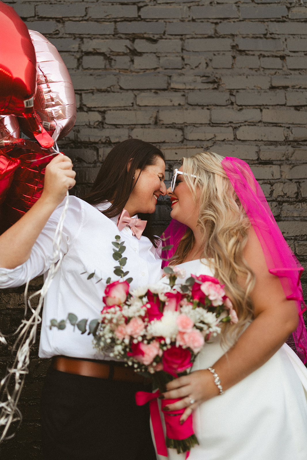 Two brides posing in front of a black brick wall with pink and red heart balloons showing off tons of unique wedding ideas